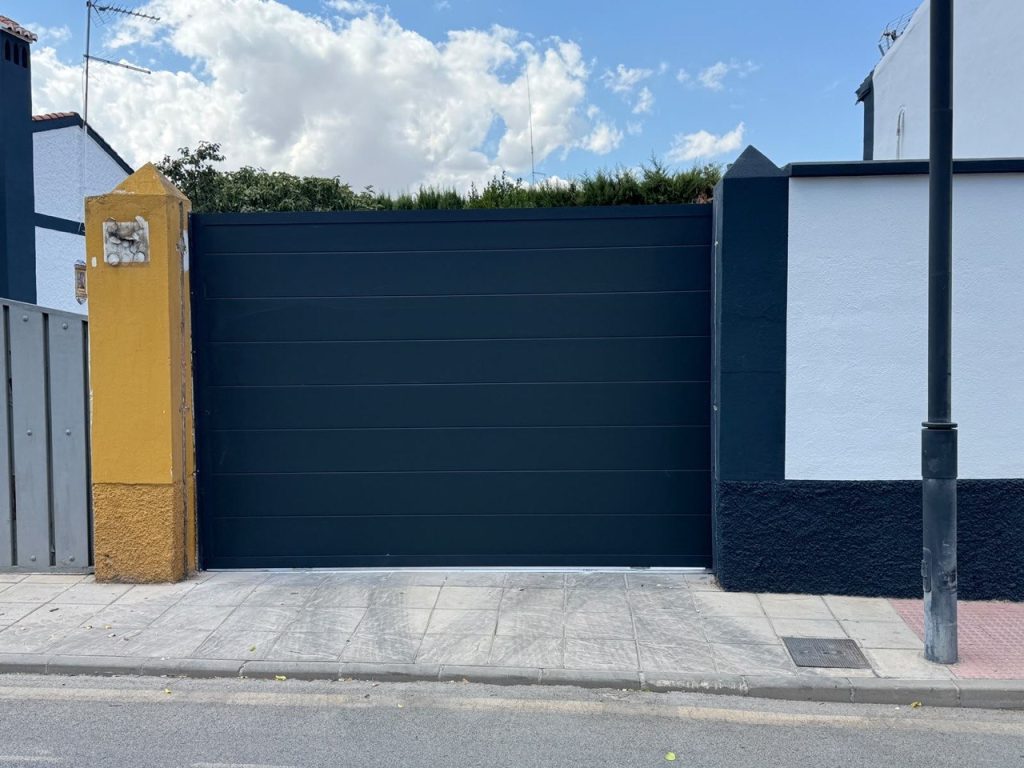 Puerta corredera automática de panel de 40 mm con acanalado horizontal en tono oscuro, cerrada a ras de acera en Híjar (Granada)